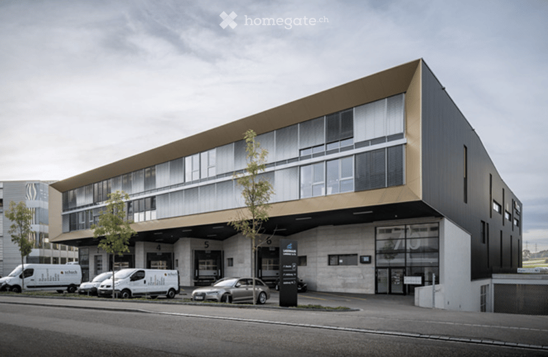 Modern multi-story building with large windows and a sleek design, featuring a parking area with several vans and a car.