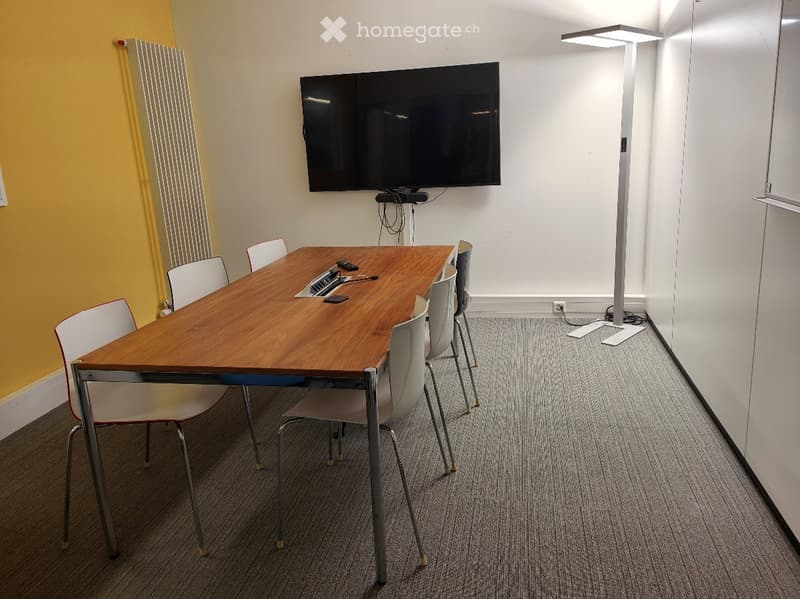 A modern meeting room with a wooden table, white chairs, a large wall-mounted screen, and a sleek floor lamp.