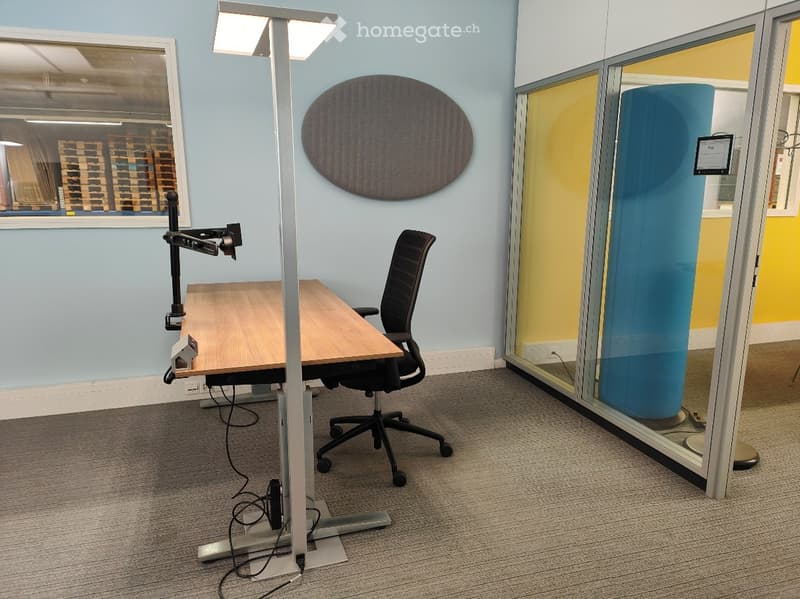 Modern office space with a wooden desk, ergonomic chair, adjustable desk lamp, and a large screen on the wall. The room features light blue walls, a glass partition, and a grey carpeted floor.