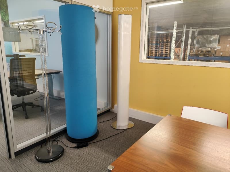 Office space with a grey carpet, a black office chair, a wooden table, a coat rack, and two large cylindrical objects (one blue, one white) in the center.