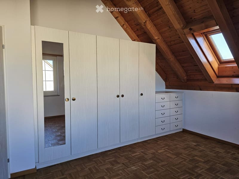 Empty attic room with wooden slanted ceilings, white built-in wardrobes, and a small window. The floor is made of herringbone-patterned wood.