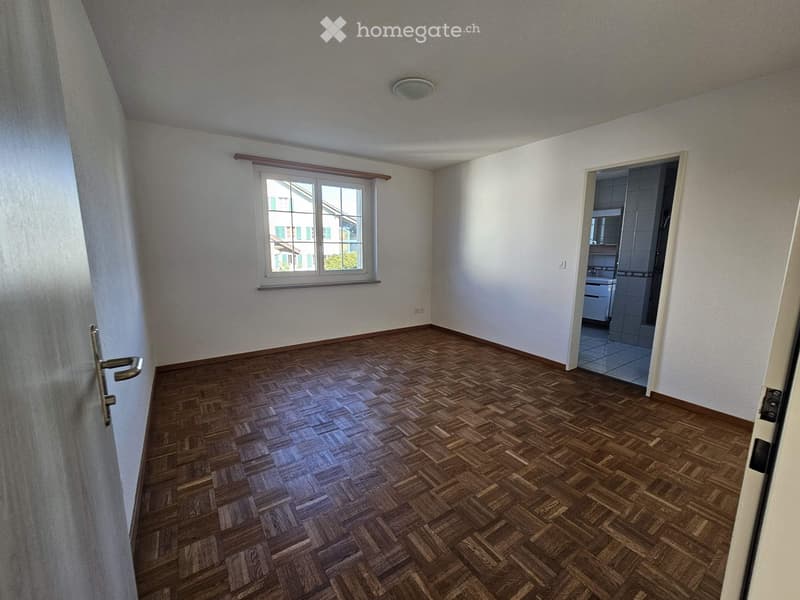 Empty room with wooden herringbone flooring, white walls, and a window with a curtain. An open doorway leads to a tiled bathroom area.