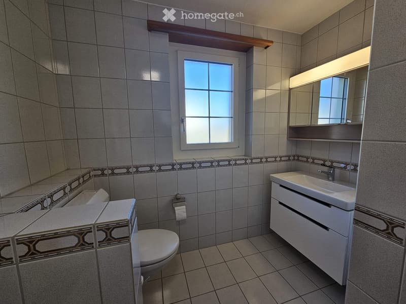 Modern bathroom with tiled walls and floor, a toilet, a sink with a mirror, and a window for natural light.