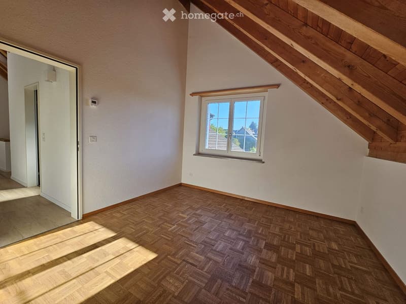Empty attic room with wooden herringbone flooring, white walls, and a single window offering a view of the outside. The room features exposed wooden beams on the ceiling and a small open doorway leading to another area.