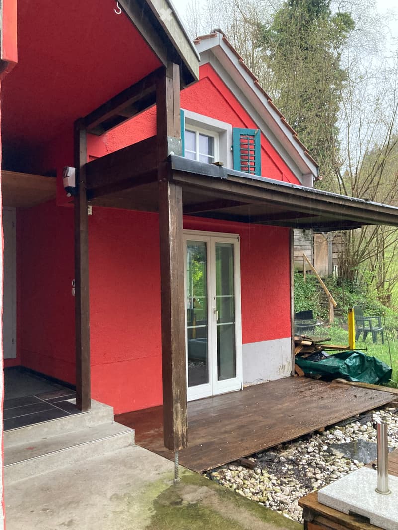 Red two-story house with a wooden deck, raised ground floor, and surrounding greenery.