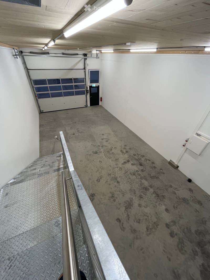 Empty concrete corridor with a metal staircase leading up, white walls, and a large garage door at the end.