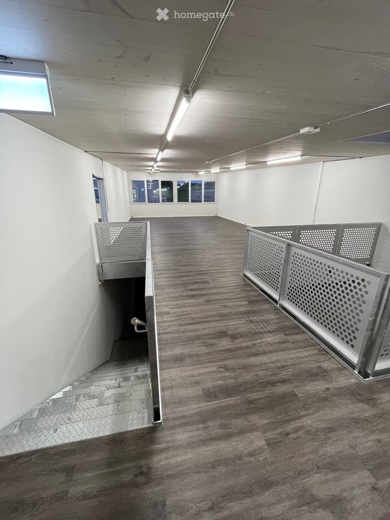 Empty hallway with wooden flooring, white walls, and a staircase leading to another level. The area is well-lit with ceiling lights and has a metal railing on one side.
