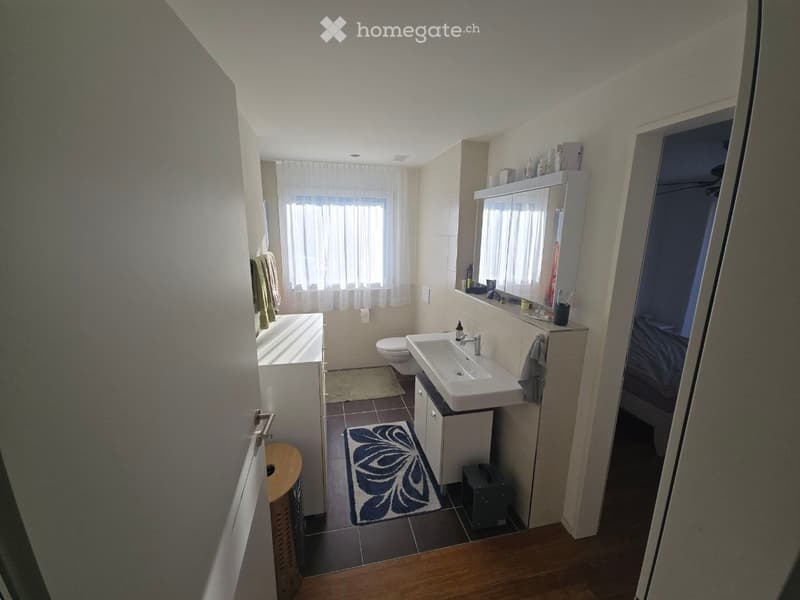 Modern bathroom with a bathtub, toilet, sink, mirror, and cabinet. Light-colored tiles and wooden flooring are visible, along with towels and a decorative rug.