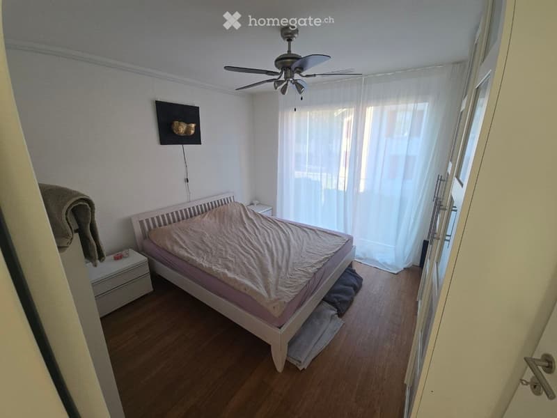 Bedroom with wooden floor, white walls, a bed with a beige duvet, a nightstand, a ceiling fan, and a large window with white curtains.