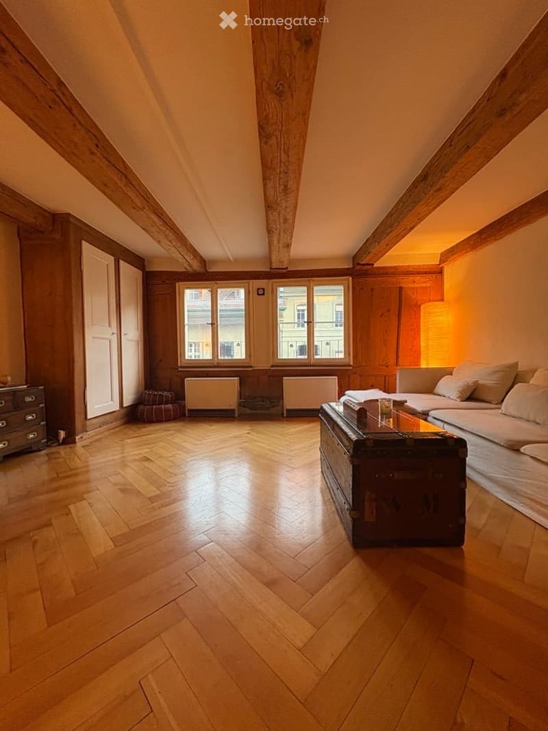 Bright living room with wooden herringbone flooring, exposed wooden beams, large windows, sofa, coffee table, chest, lamp, and a small stool.