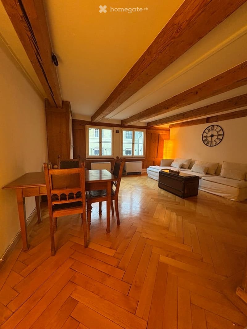 Bright, open room with wooden floors and exposed wooden beams. Features a sofa, a small table with chairs, and a wall-mounted clock.