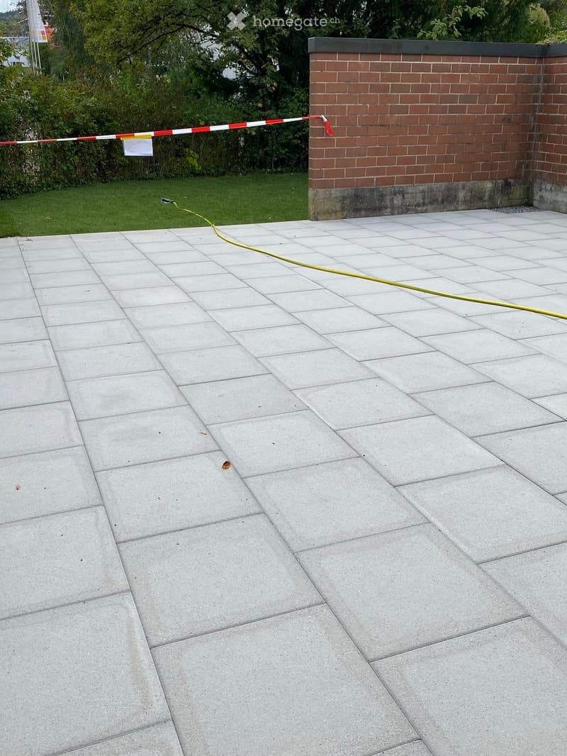 A paved outdoor area with large concrete tiles, a brick wall, and a grassy area with trees in the background.