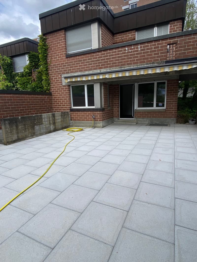 Patio area with large concrete tiles, yellow hose, and part of a brick house with windows and a covered entrance.