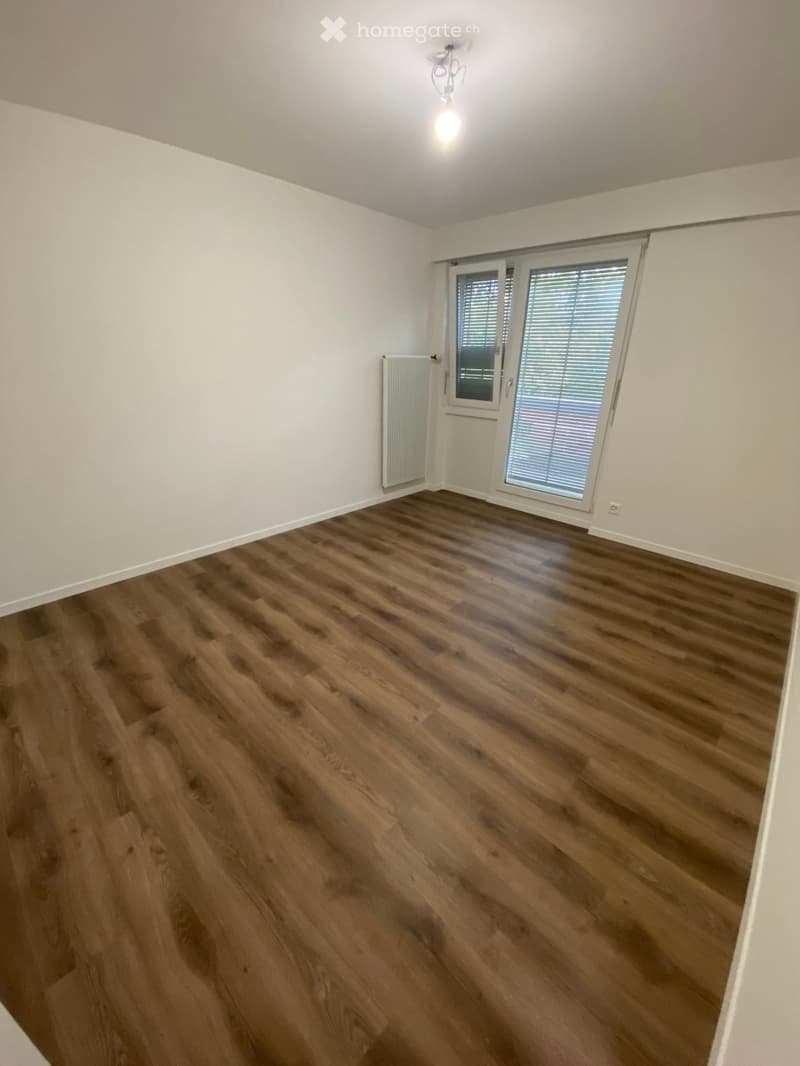 Empty room with wooden flooring, white walls, and a large window with blinds. A radiator is visible on one wall.