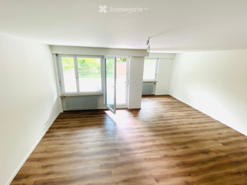 Empty room with wooden flooring, white walls, large windows, and a glass door leading outside.