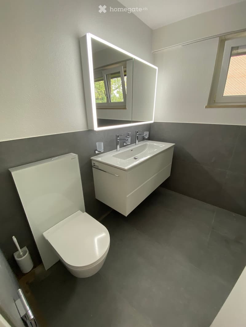 Modern bathroom with a white toilet, a white sink with a modern faucet, a large illuminated mirror, and a dark grey tiled floor.