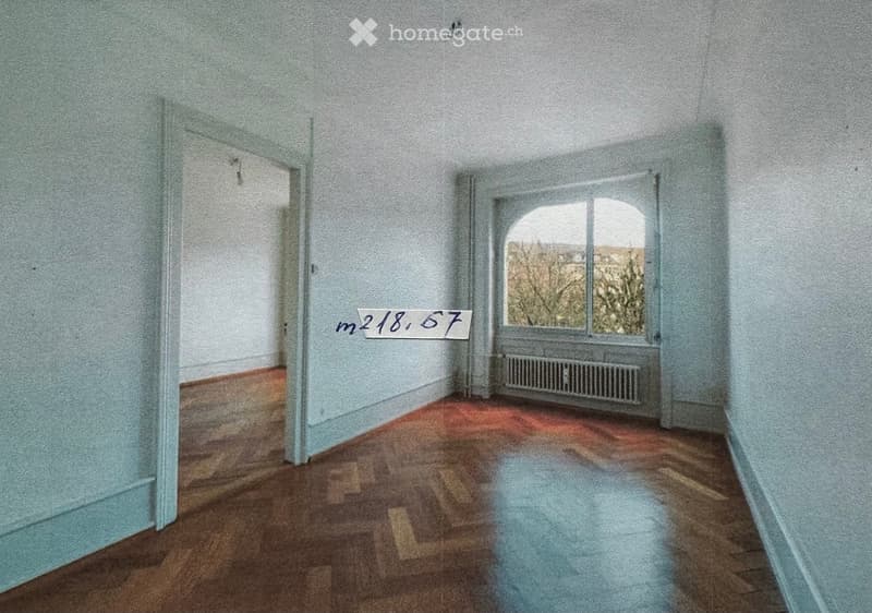 Empty room with wooden flooring, light blue walls, and a large window with a radiator below it. The room is completely unfurnished.