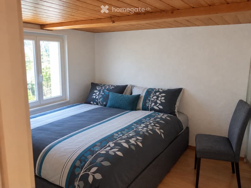 Cozy bedroom with a double bed, blue and white patterned bedding, and a matching chair. The room features light wooden flooring and white walls, with a window offering a view of greenery outside.