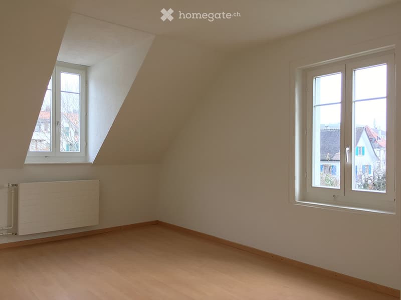 Empty attic room with wooden flooring, white walls, and two windows, one with a slanted roofline. A radiator is visible on the left wall.