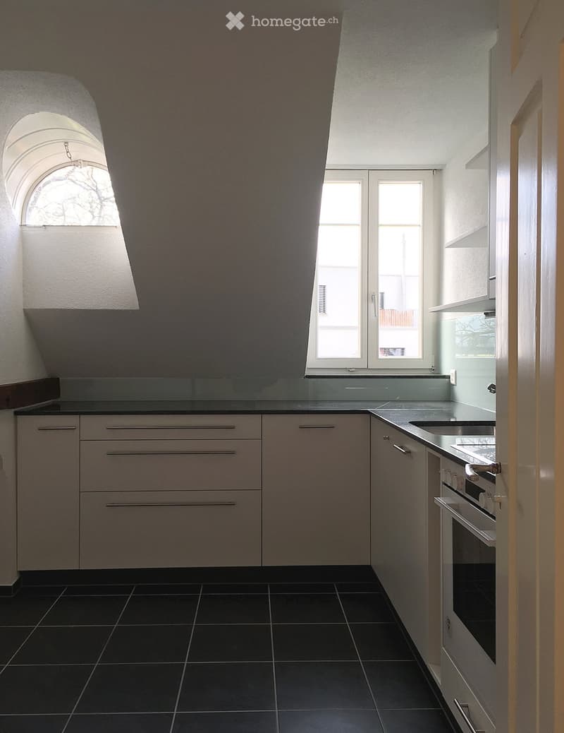 Modern kitchen with white cabinets, black tiled floor, and stainless steel appliances including a sink and oven. Features large windows and an arched window on the left side.