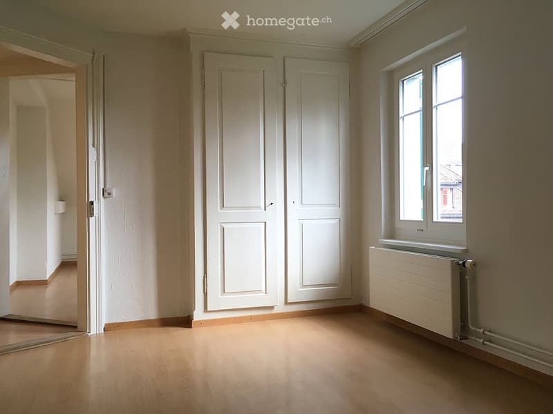 Empty room with wooden flooring, white walls, and a window with a radiator below it. Two white doors are visible on the wall.