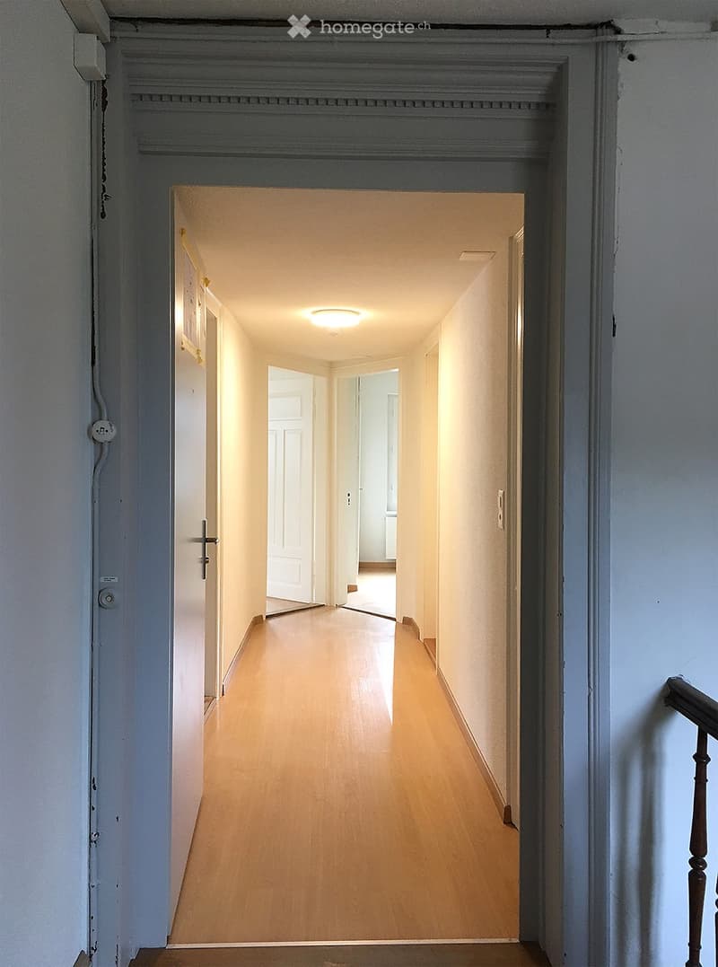 Empty hallway with wooden flooring, white walls, and recessed lighting. Doors lead to other rooms, and a staircase is visible on the right side.