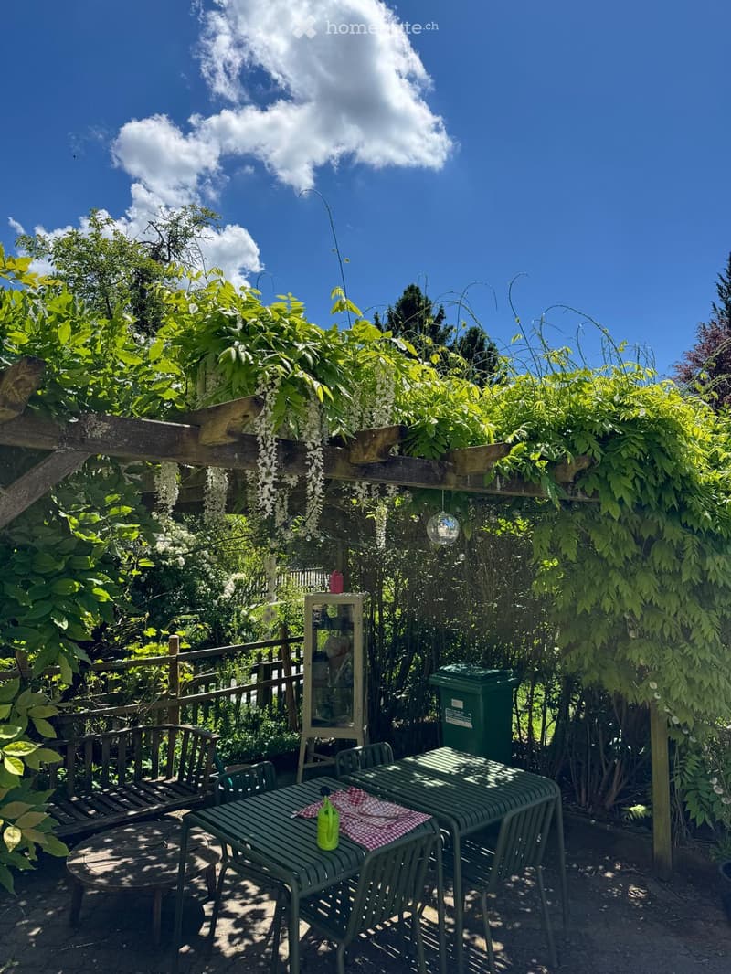 A lush garden area with a wooden pergola, green metal table and chairs, a bench, and a small mirror on a stand.