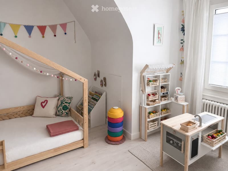A cozy children's bedroom with a wooden bed, a small play kitchen area, and a shelf filled with toys and books. The room features a slanted ceiling and large windows with white curtains.