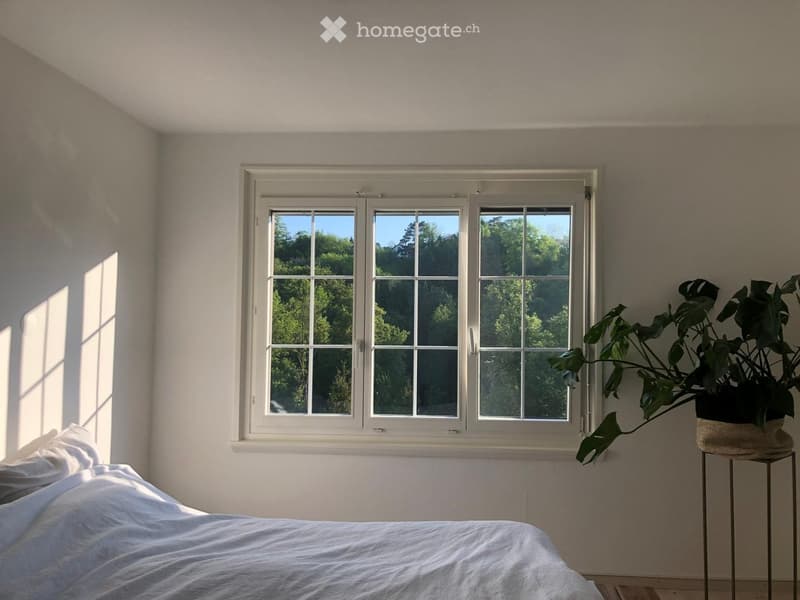 Bright bedroom with a large window showing greenery outside, a neatly made bed, and a potted plant on a small stool.