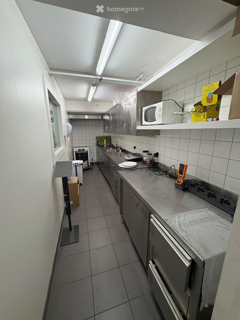 Modern kitchen with stainless steel appliances, tiled walls, and a grey countertop. Includes a microwave, oven, refrigerator, and various kitchen items on the counter.
