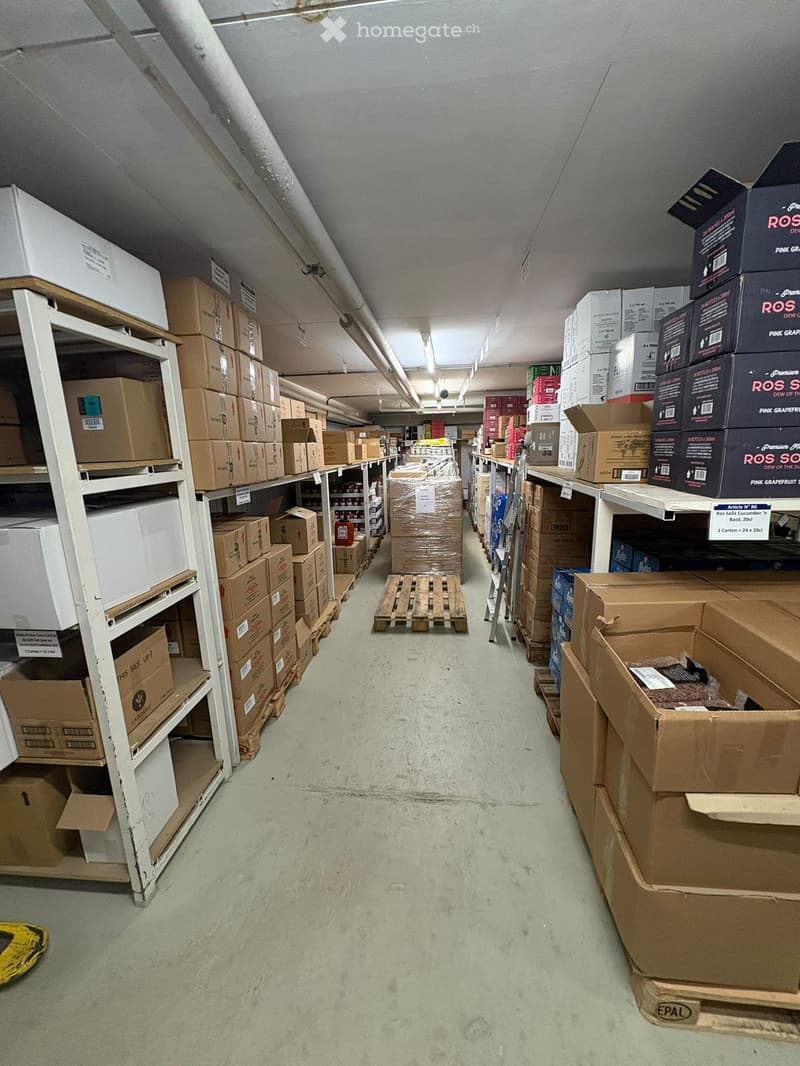 A long, narrow storage corridor with metal shelving units on both sides, filled with cardboard boxes and pallets. The ceiling has exposed pipes and fluorescent lighting.