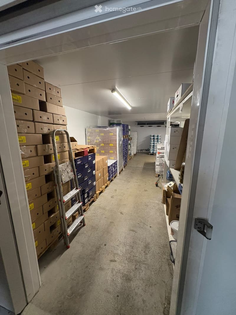 A narrow, well-lit storage corridor with concrete floors and white walls, lined with shelves and stacked cardboard boxes on both sides.