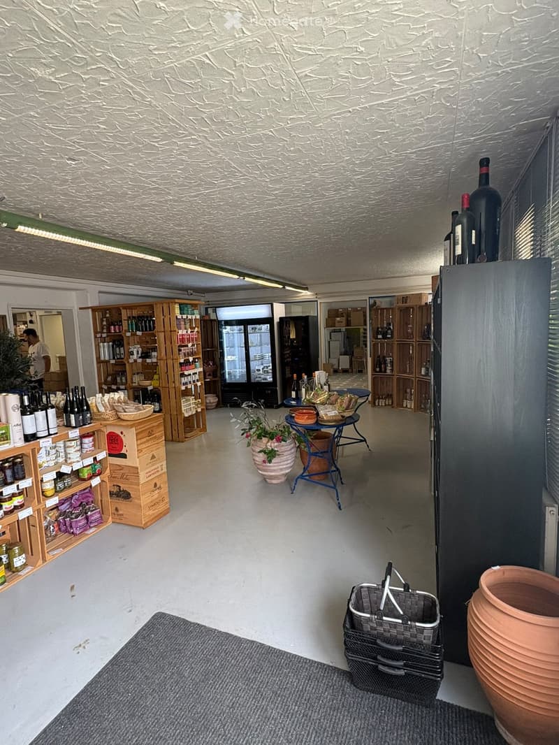 A well-lit store interior with wooden shelves stocked with various products, including wine bottles and jars. A small round table with a potted plant sits in the center, and a shopping basket is placed near the entrance.