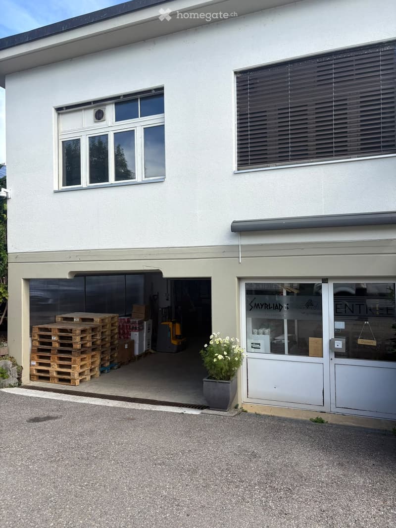 Two-story building with a garage entrance, stacked pallets, and a potted plant near the entrance.