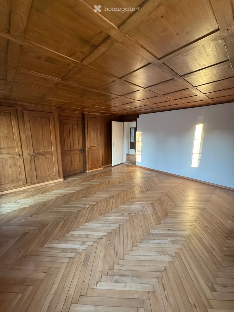 Empty room with wooden herringbone flooring, exposed wooden ceiling beams, and white walls. Features include wooden doors and a small adjacent space visible through an open doorway.