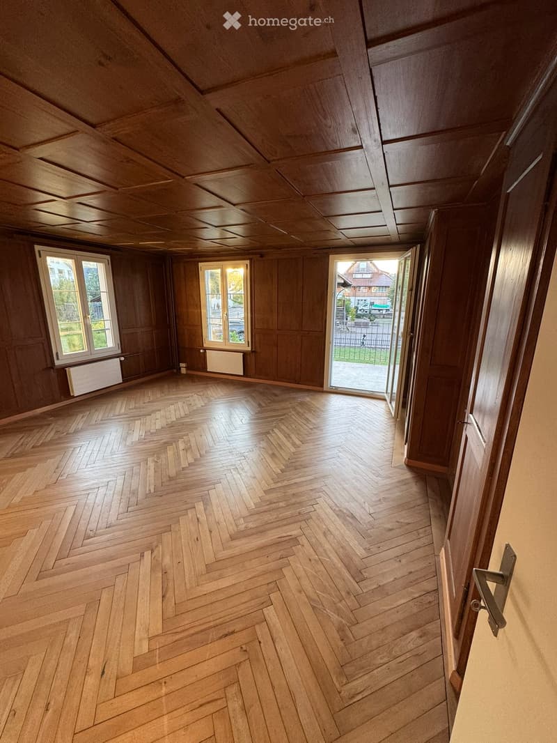 Empty room with wooden herringbone flooring, wooden walls, and large windows, featuring a door leading outside.