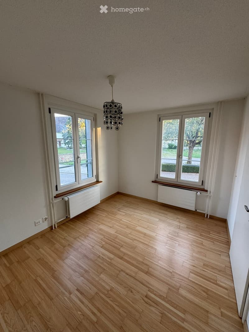 Empty room with wooden flooring, large windows, radiator, and a modern chandelier.