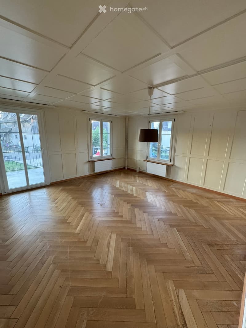 Empty room with wooden herringbone flooring, white walls, large windows, and a hanging light fixture.