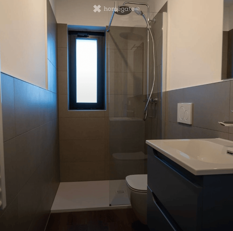 Modern bathroom with a glass-enclosed shower, toilet, sink, and a window providing natural light.