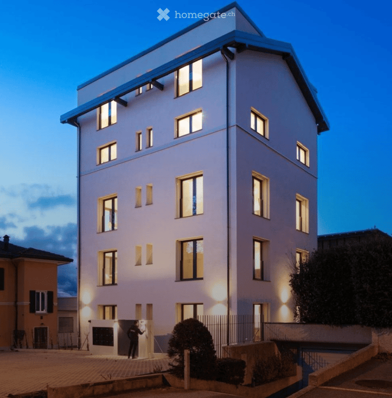 Modern multi-story residential building with illuminated windows and clean lines, featuring a raised ground floor and contemporary design.