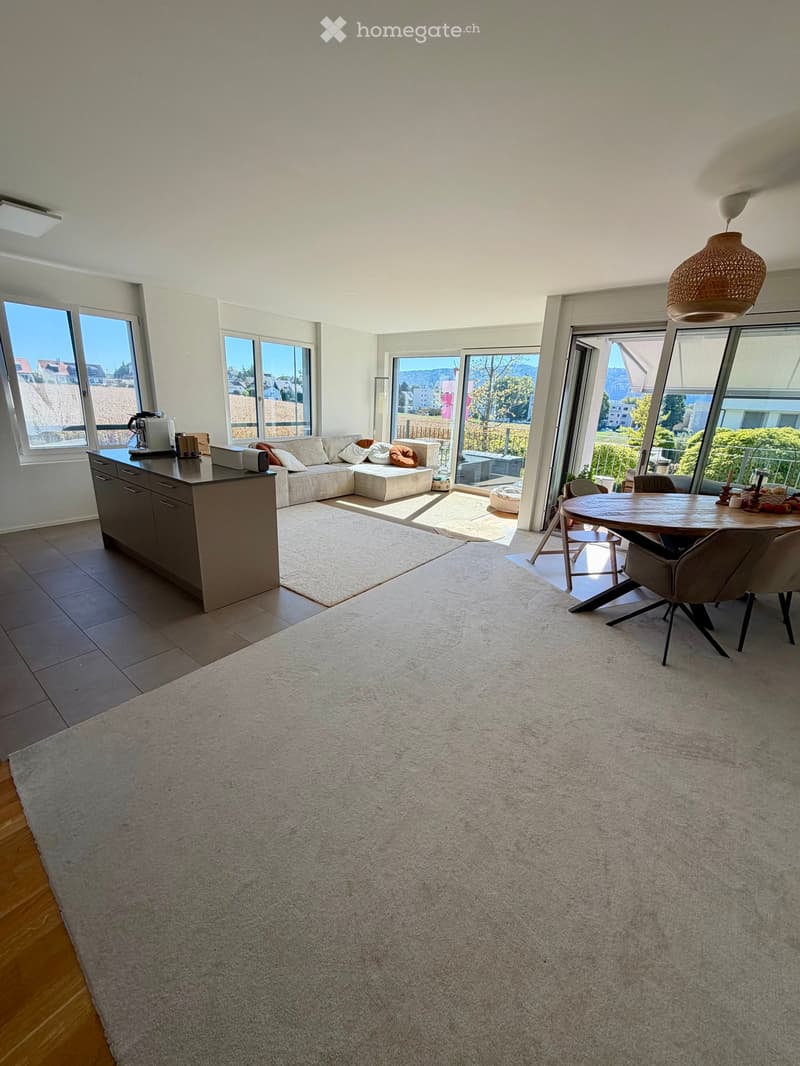 Bright, open living and dining area with large windows offering a scenic view, featuring a sectional sofa, coffee table, kitchen island, dining table, and pendant light.