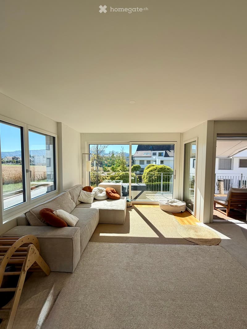 Modern living room with large windows, a sectional sofa with colorful pillows, a wooden chair, a small table, and a floor lamp. The room features a carpeted floor and opens to a balcony with outdoor seating.