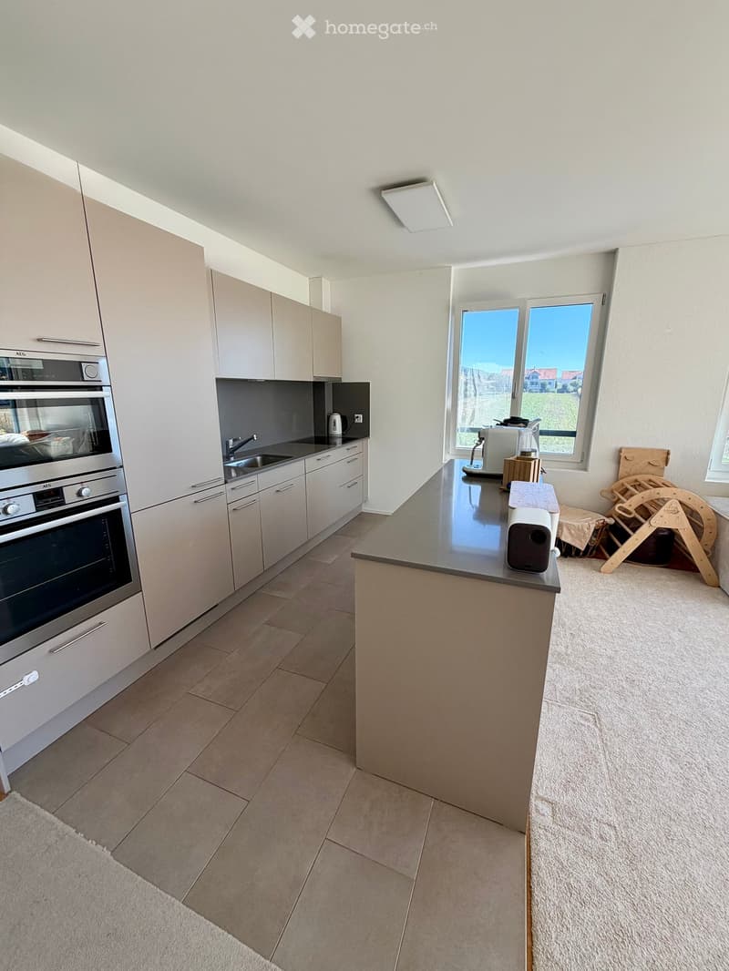 Modern kitchen with light-colored cabinets, stainless steel oven, sink, and countertop. Features a central island with a coffee maker and blender, and a speaker on the counter.