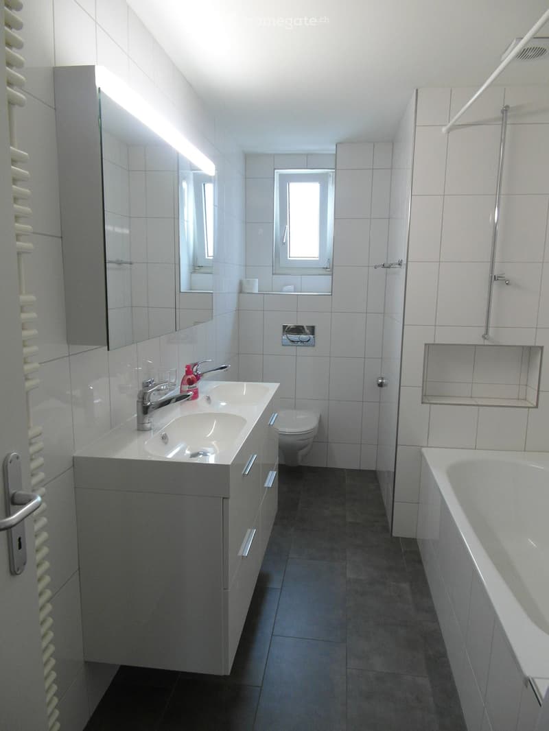 Modern bathroom with white tiles, a double sink, toilet, bathtub, and a large mirror above the sink. The room features a radiator on the left and a window above the toilet.