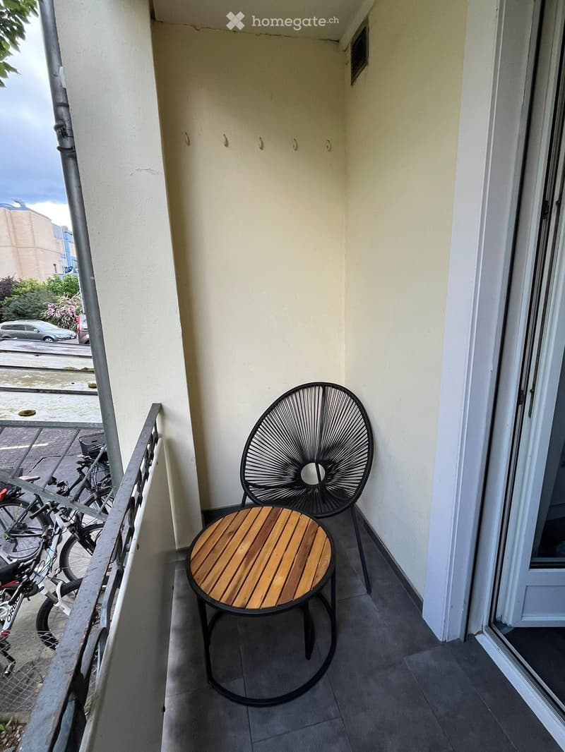 Balcony with a small wooden table and a black wire chair, overlooking a street with parked cars and bicycles.