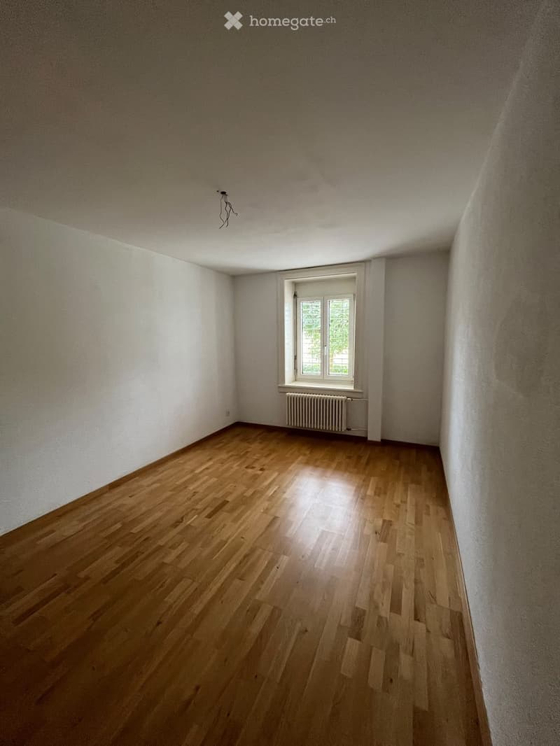 Empty room with wooden flooring, white walls, and a single window with a radiator below it.