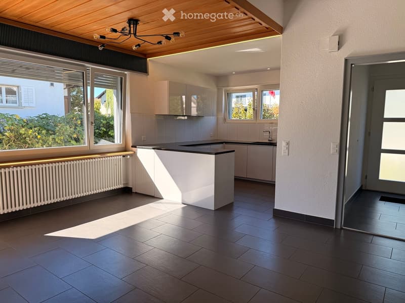 Modern kitchen with dark wooden flooring, white cabinets, black countertop, and large windows allowing natural light.