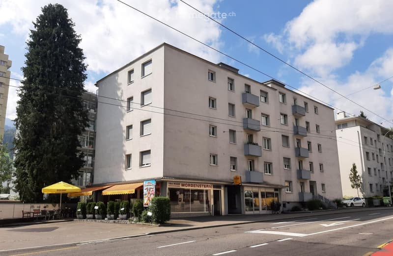 A multi-story apartment building with a cafe or restaurant on the ground floor. The building has a beige exterior and several balconies. There is a large tree in front of the building and power lines running overhead.