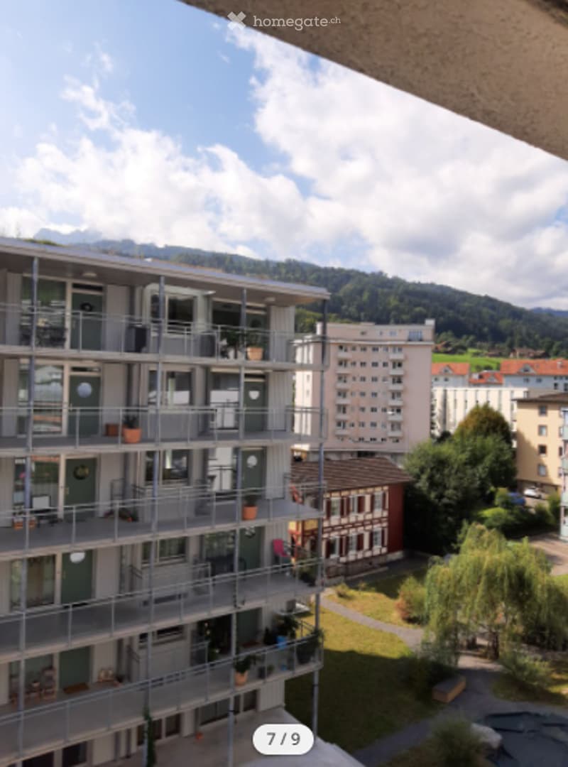 The building is a multi-story residential apartment complex with balconies. In the foreground, there is a walkway leading to a lawn area with plants and trees. There are mountains in the background. It is the 7th image out of 9.