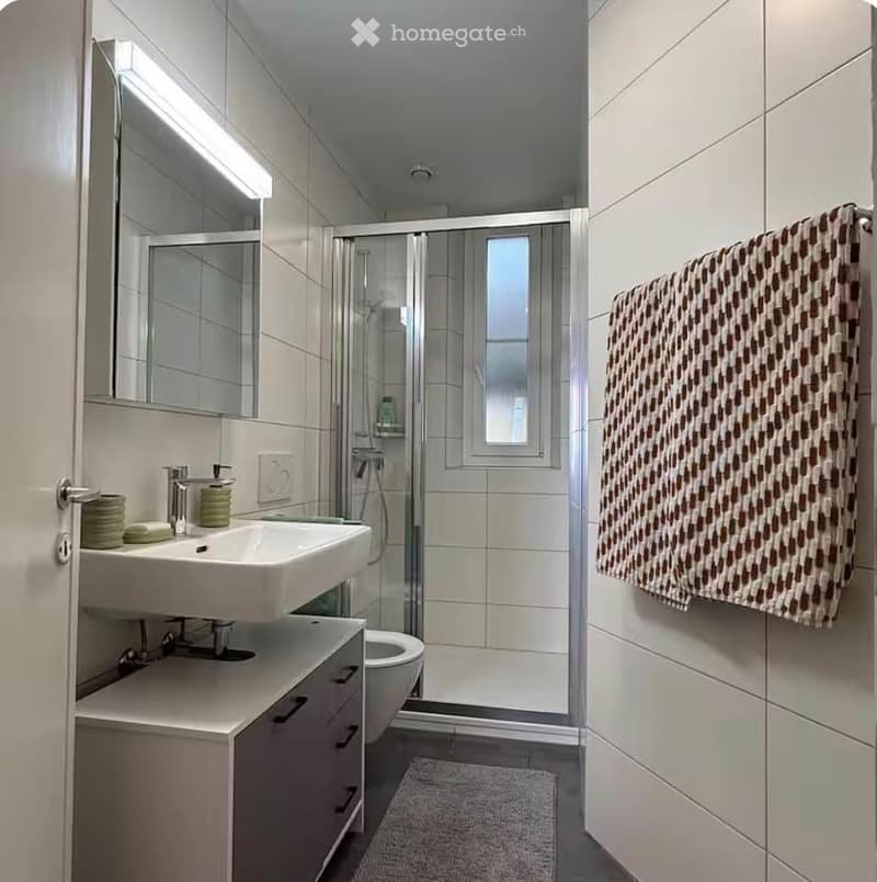 Modern bathroom with white tiles, a double sink, a large mirror, a shower, and a towel hanging on the wall.